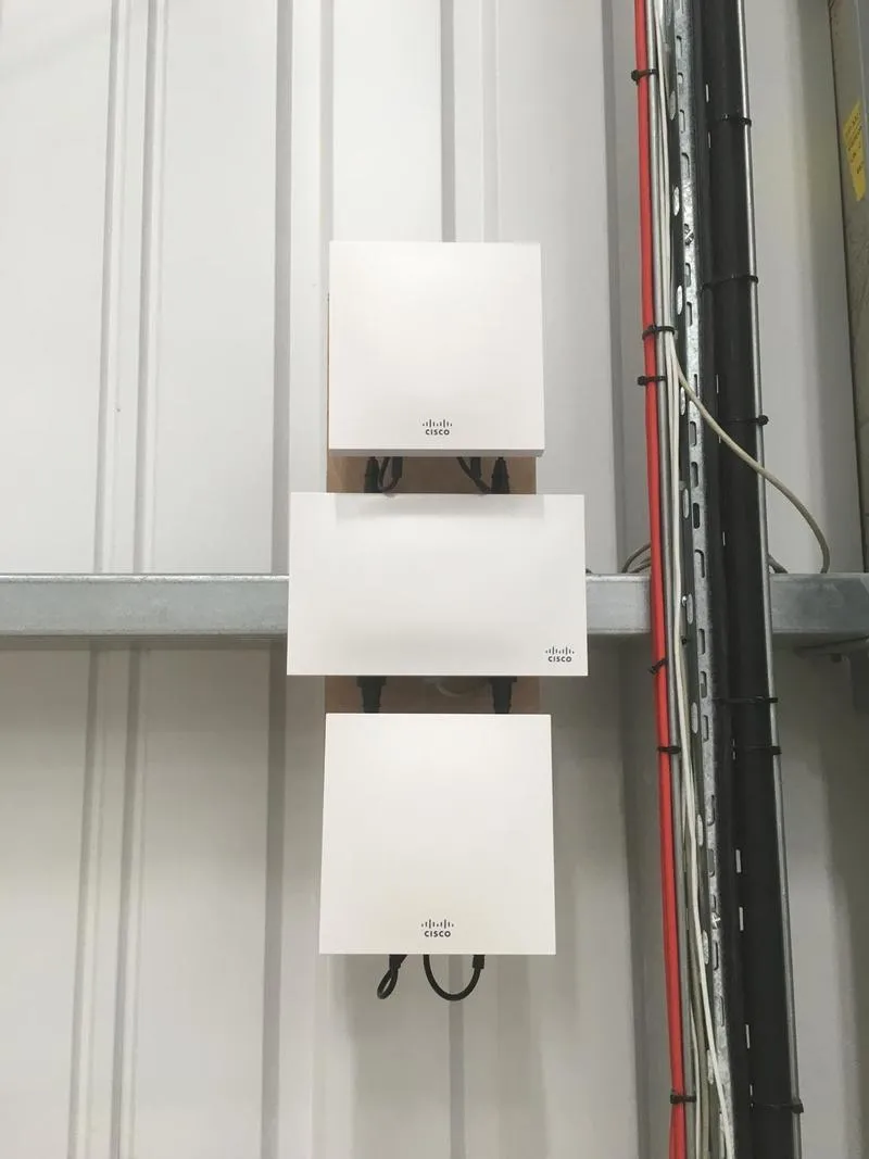 Three access points mounted on timber backboards fixed to a profiled steel wall in an industrial unit, with containment and cabling beside them