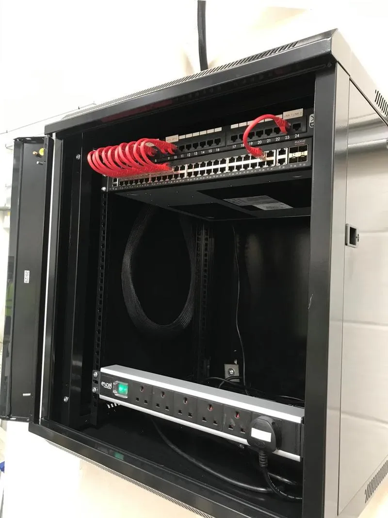 An open wall cabinet with a Ruckus switch and a patch panel whose ports are labelled for individual access points