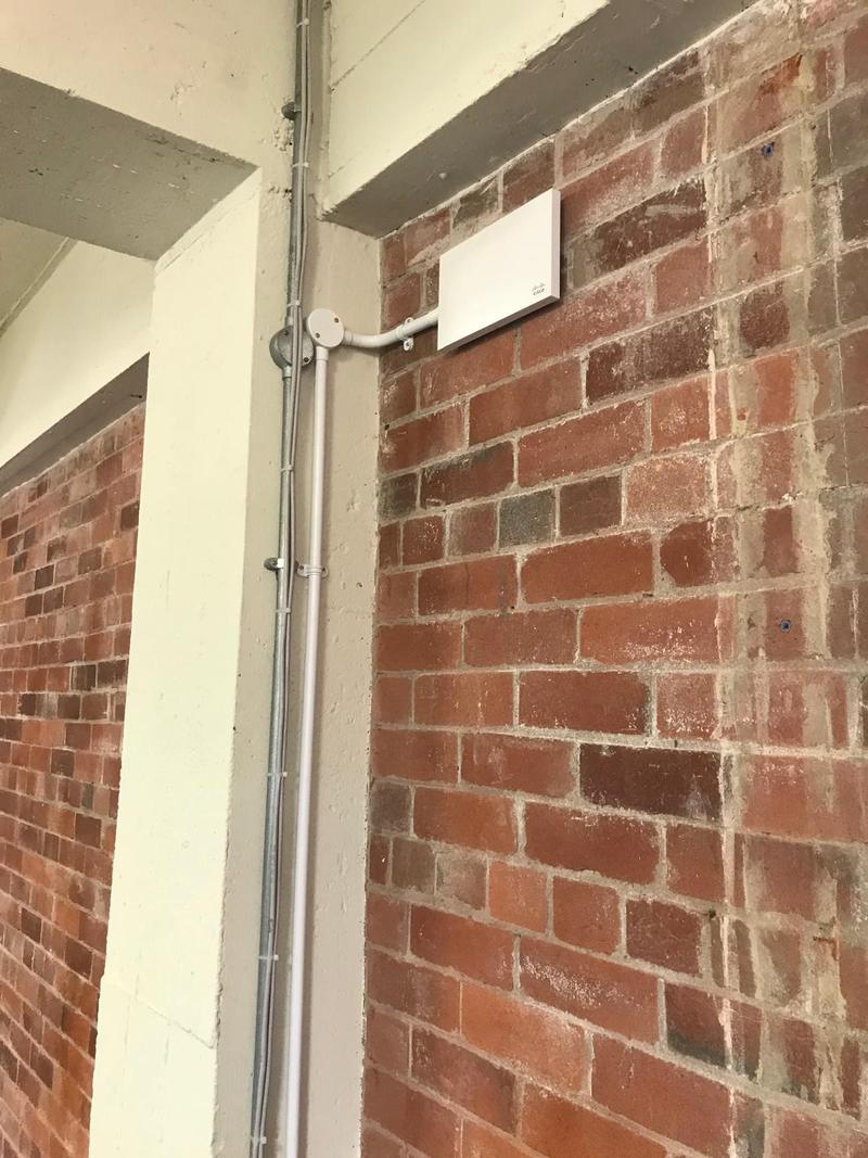 A white Cisco access point mounted on an exposed brick wall high under a concrete beam, fed by a white conduit dropper run neatly down the corner from the containment above