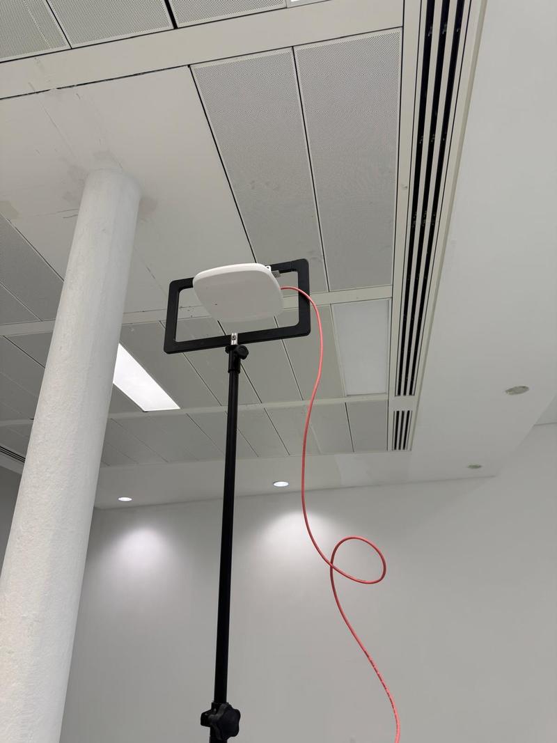 A close view of an access point clamped in a survey bracket just below a suspended ceiling grid, a red patch lead running from it