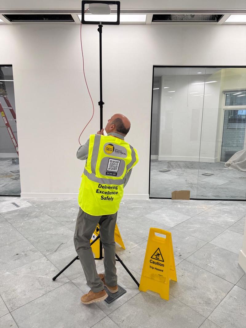 An engineer in SEG branded hi-vis reaching up to position an access point against an office ceiling, with trip hazard signs placed around the working area
