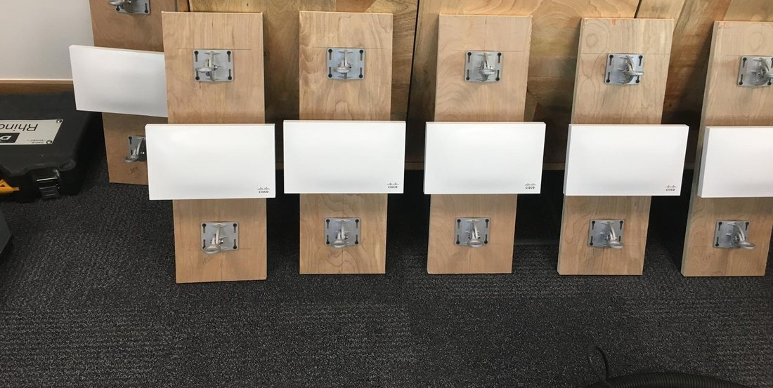 Six access points and their mounting brackets prepared on timber backboards and stood in a row on the floor before installation, with cable glands laid out on the side unit behind