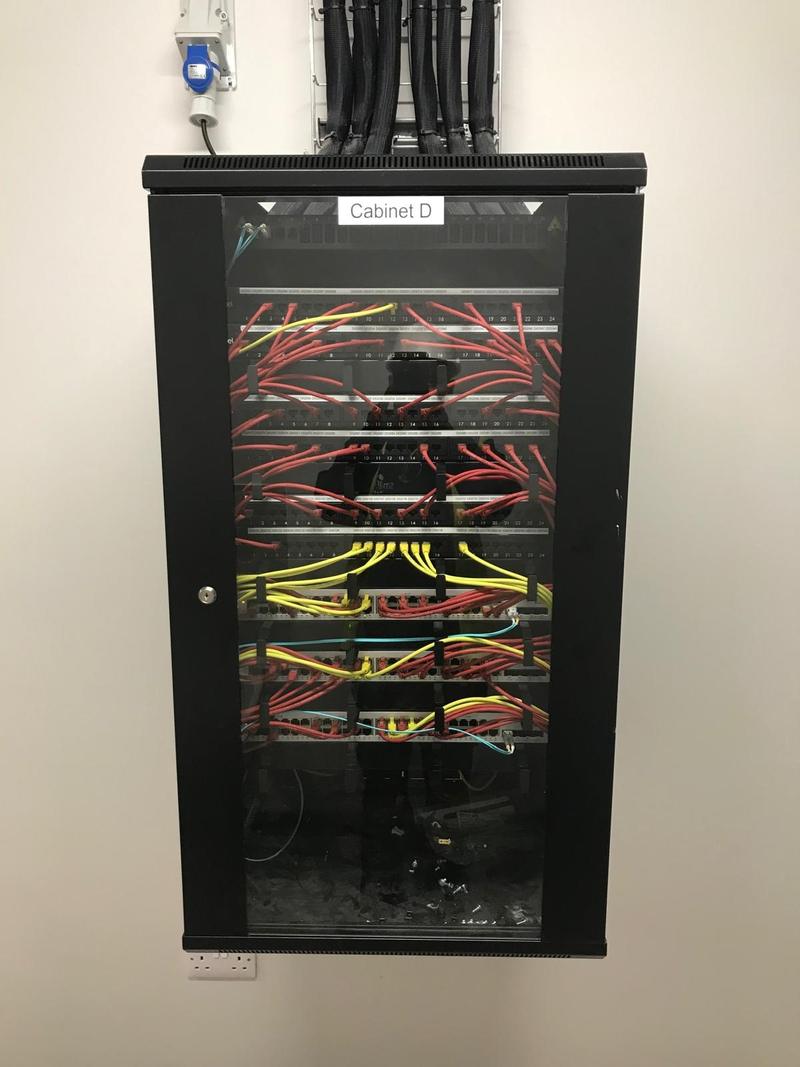A glass-fronted wall cabinet labelled Cabinet D, with four labelled patch panels patched out in red and yellow leads, every lead dressed through the horizontal management bars
