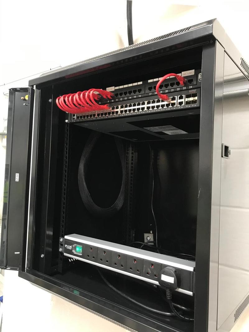 An open wall cabinet with a Ruckus switch, a patch panel whose first ports are labelled AP 9 and AP 10, red patch leads looped between them and a rack PDU below