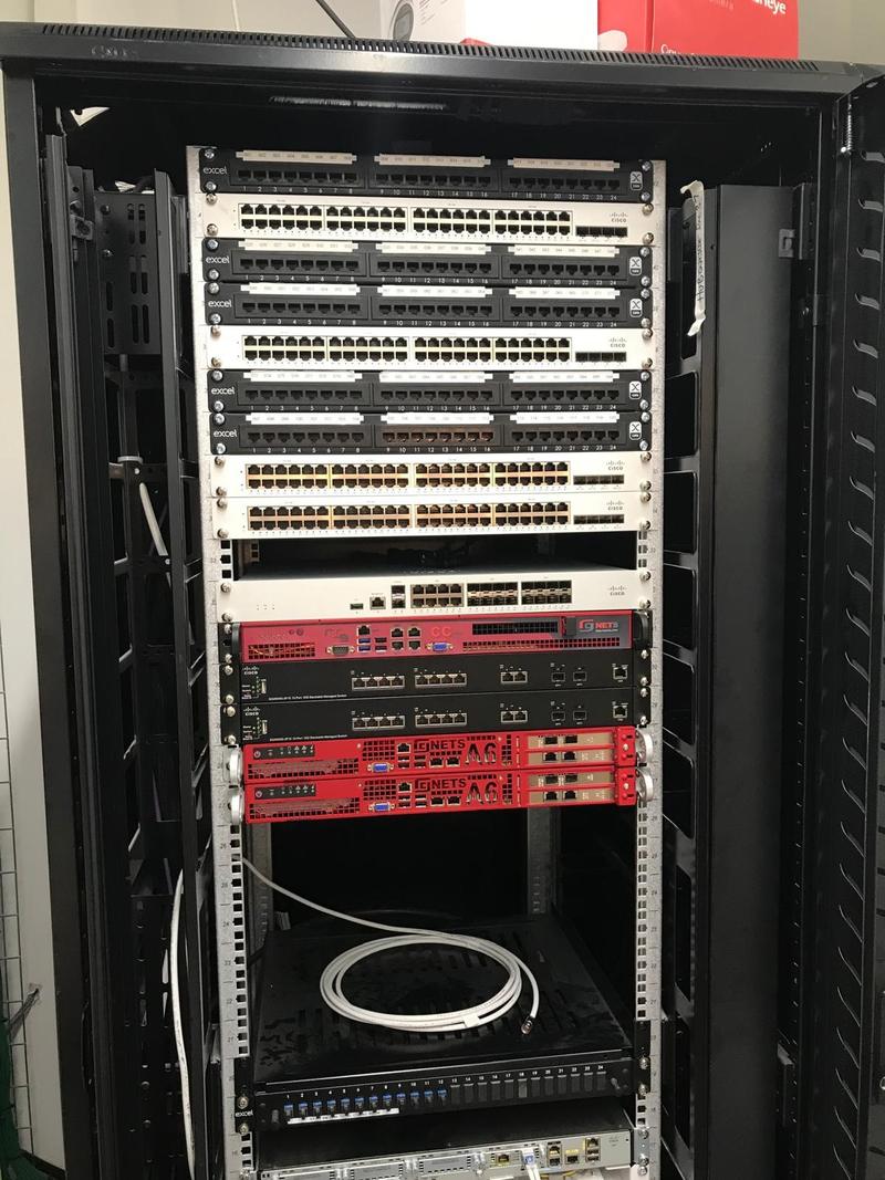 A full-height open comms rack with six labelled Excel patch panels, four Cisco switches, a core switch and two red rack-mounted security appliances, each item screwed to its own U position