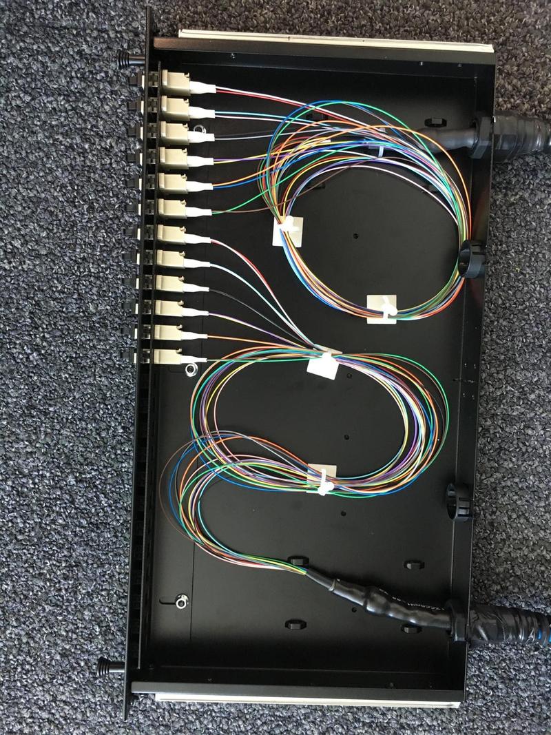 An open fibre patch panel tray with colour-coded pigtails coiled inside it and terminated into a row of adapters, the incoming cable gland at the corner