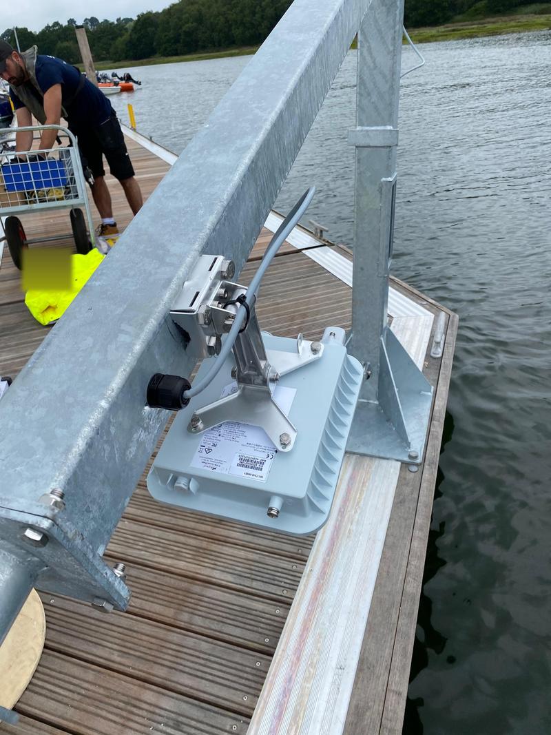 An engineer kneeling on a marina pontoon fitting an outdoor access point into a galvanised bracket on a post, with the water immediately behind