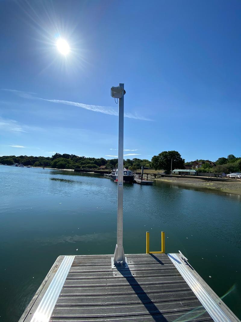 An outdoor access point on a galvanised post at the end of a marina pontoon, water and the far shore behind it