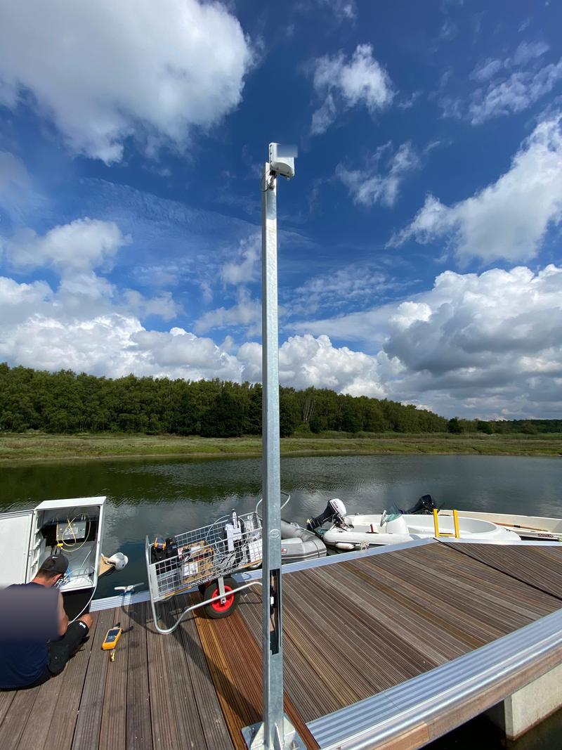 An engineer working at an open equipment enclosure on a marina pontoon, an access point on a post beside him and the water behind
