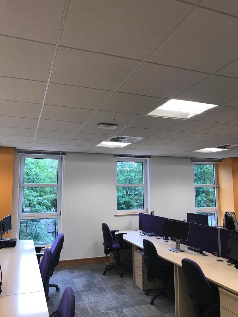 An occupied open-plan office with a suspended ceiling, recessed light fittings and air conditioning grilles, desks with screens and privacy panels along the window wall