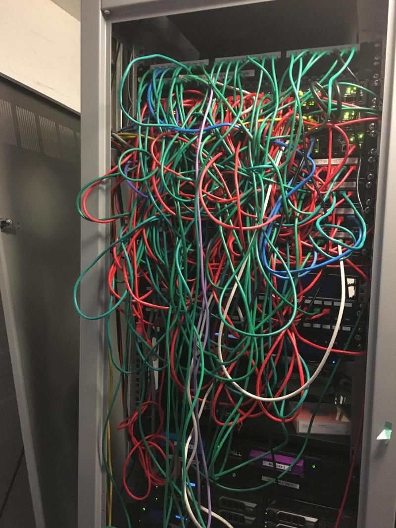 A comms cabinet with a mass of red and green patch leads hanging loose across the front of the switches, tangled together with no cable management and no discernible order
