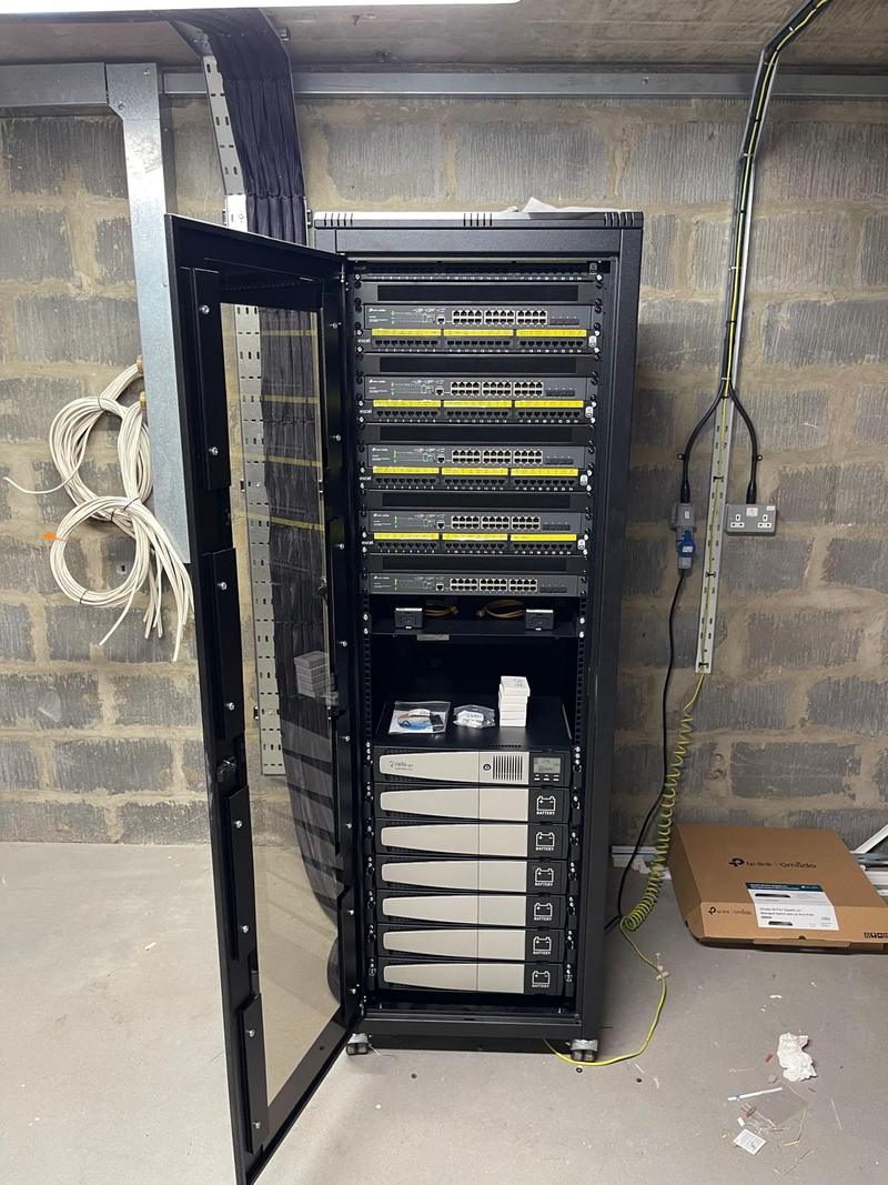 A newly built floor-standing cabinet in a plant room with four patch panels, four managed PoE switches and a rack-mounted UPS with six battery modules below them