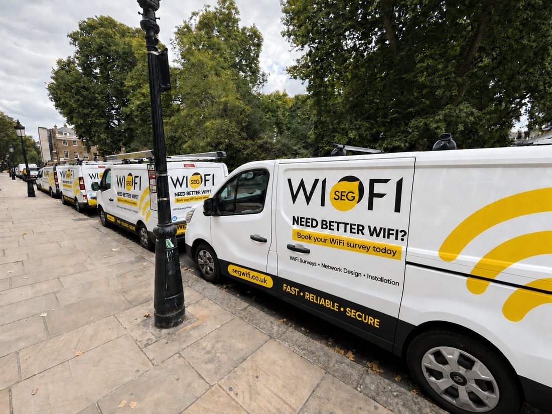 A line of SEGwifi liveried vans parked along a London street, the nearest signwritten WI-FI with the SEG roundel, “Need better WiFi? Book your WiFi survey today”, “WiFi Surveys · Network Design · Installation” and segwifi.co.uk