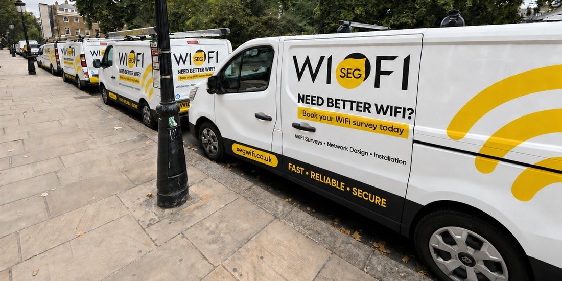 A line of SEGwifi liveried vans parked along a London street, the nearest signwritten WI-FI with the SEG roundel, “Need better WiFi? Book your WiFi survey today”, “WiFi Surveys · Network Design · Installation” and segwifi.co.uk