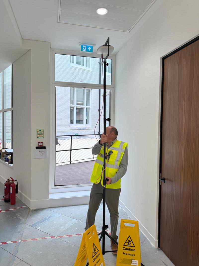 An engineer in hi-vis adjusting a survey tripod in a corridor beside a window, with trip hazard signs in the foreground