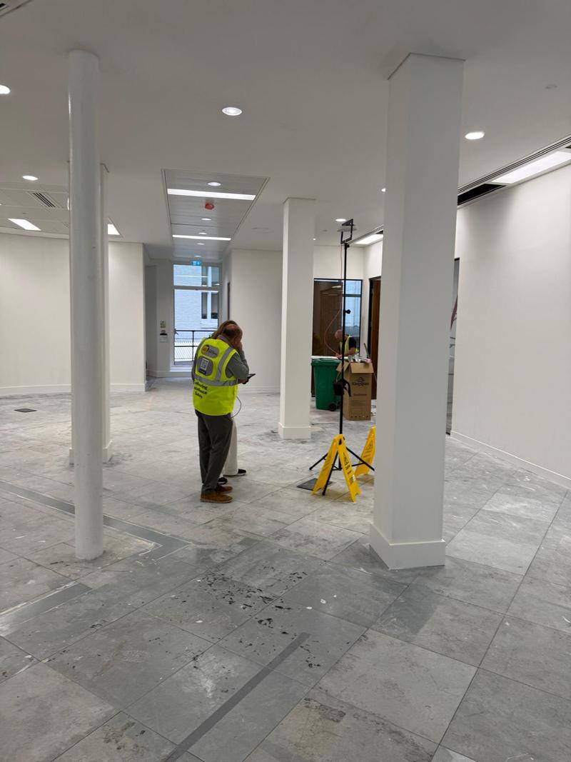 Two engineers in hi-vis working on an empty office floor with white columns, one reviewing a tablet and one at a survey tripod
