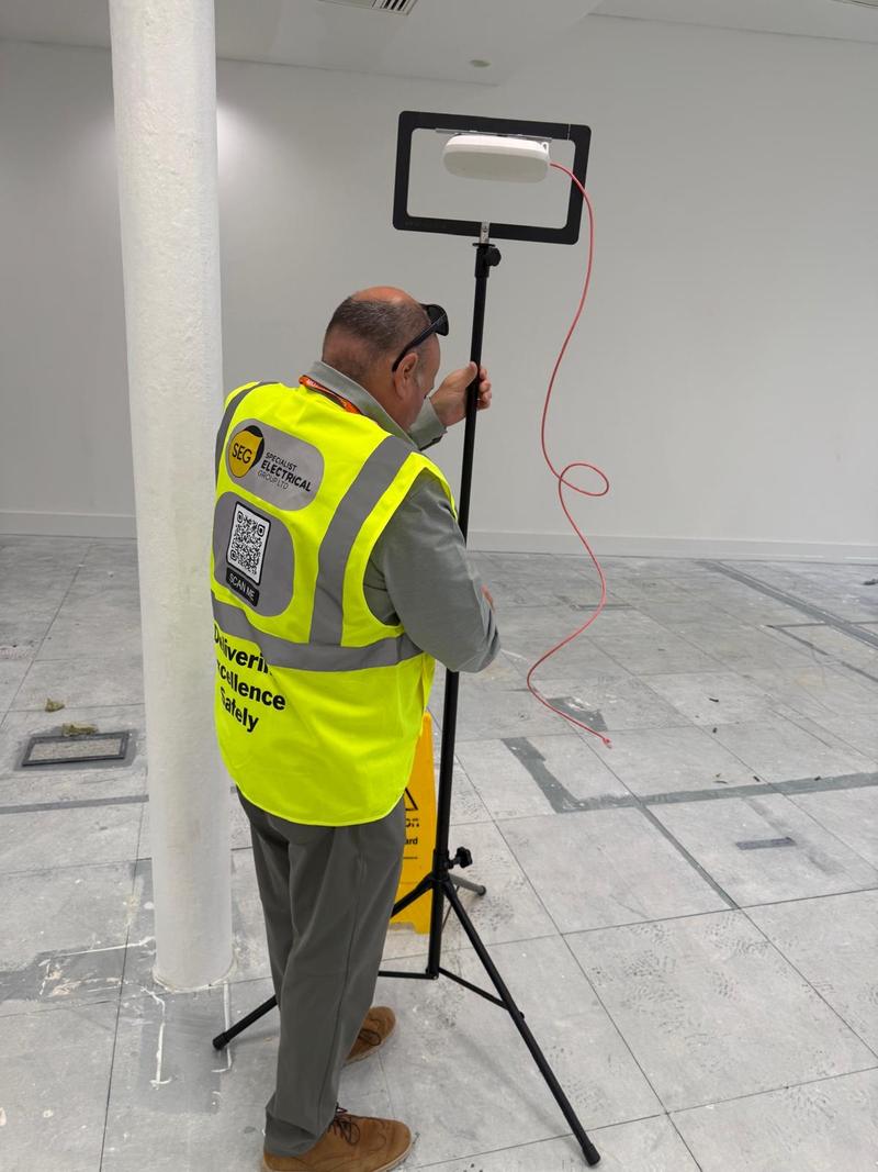 A SEGwifi engineer in branded hi-vis positioning an access point on a survey tripod beneath an office ceiling