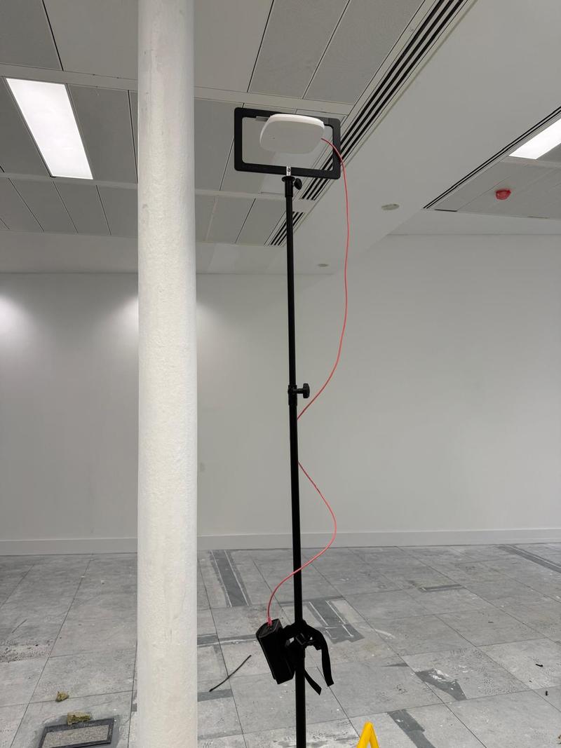 A survey tripod extended to ceiling height in an empty office fit-out, an access point on its bracket and a battery pack clamped to the mast